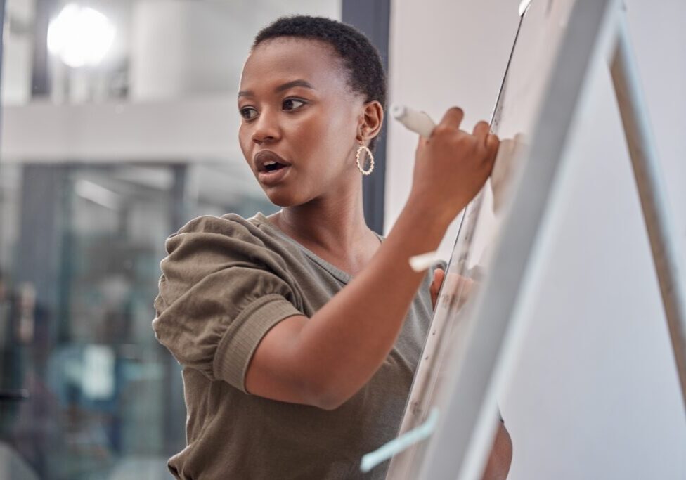 Person writing on a whiteboard while speaking, wearing earrings and a green top.