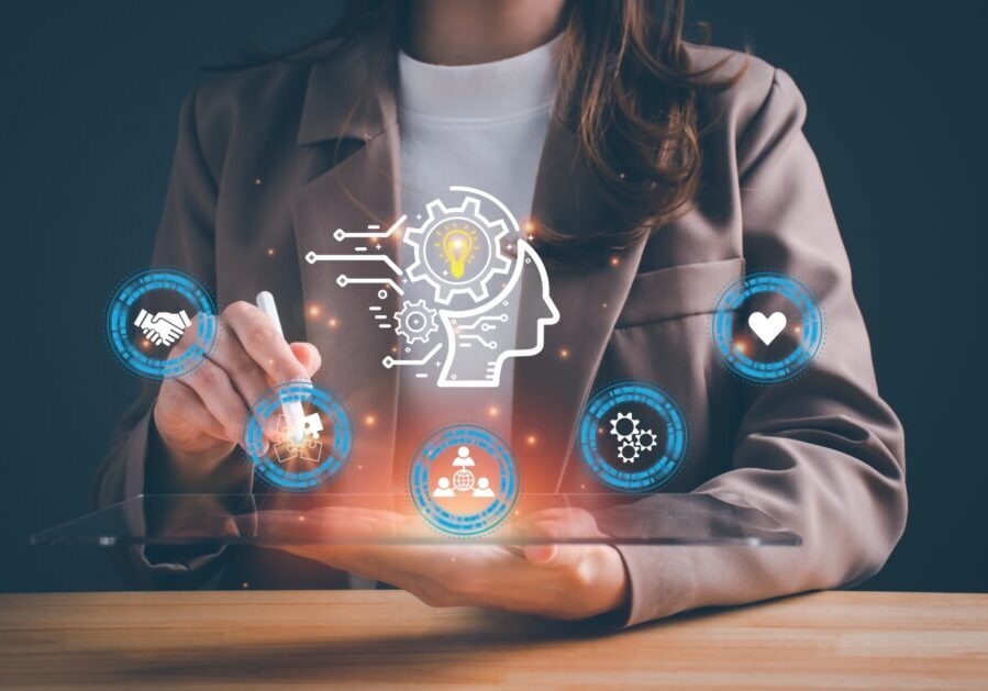 Person touching a tablet displaying futuristic digital icons including a brain with gears, handshake, gear, and lightbulb, suggesting innovation and technology.